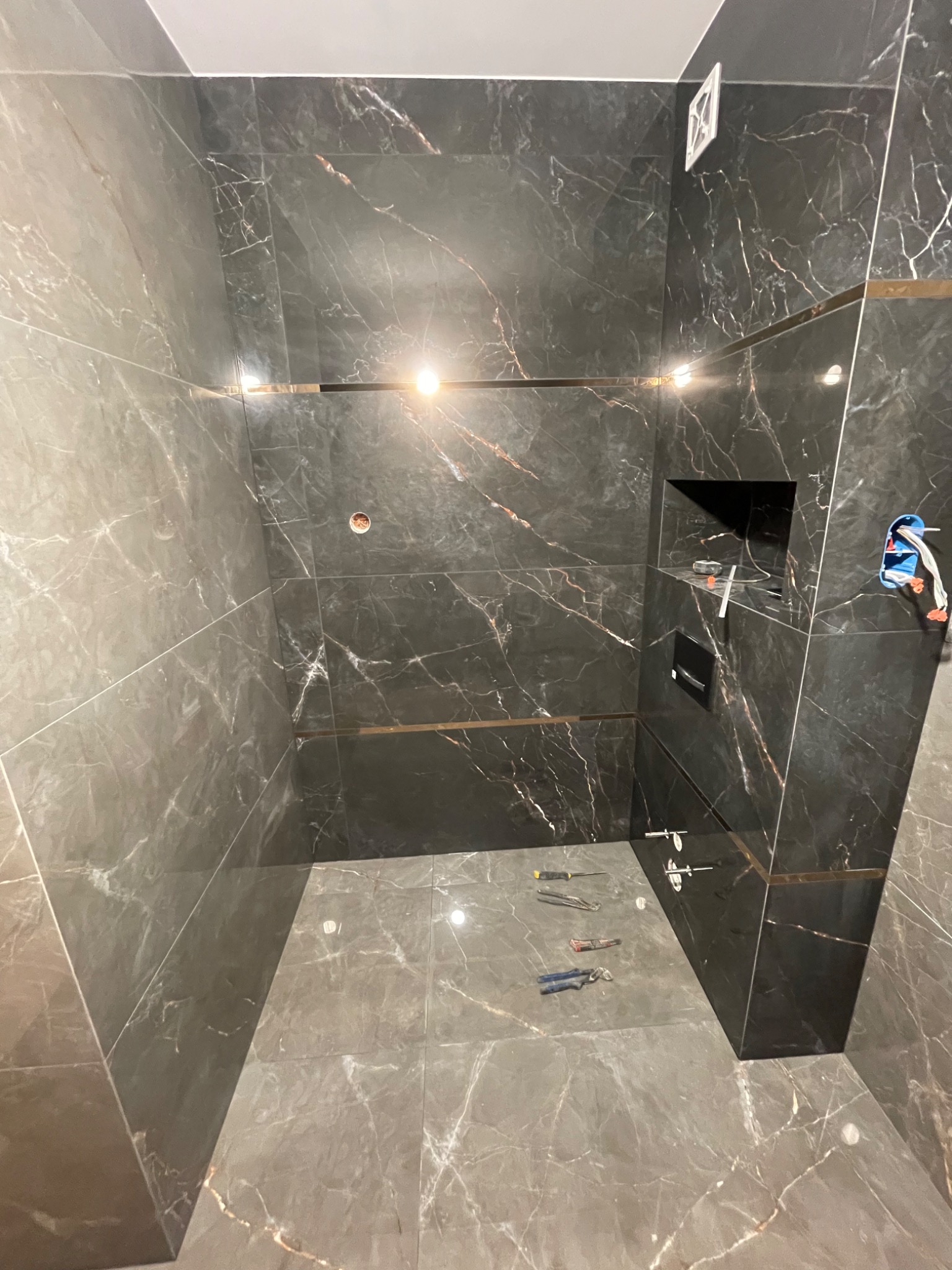 Wnętrze łazienki w trakcie wykańczania, ściany i podłoga wyłożone ciemnymi płytkami imitującymi marmur z delikatnymi złotymi akcentami, widoczne narzędzia glazurnicze na podłodze, otwór...