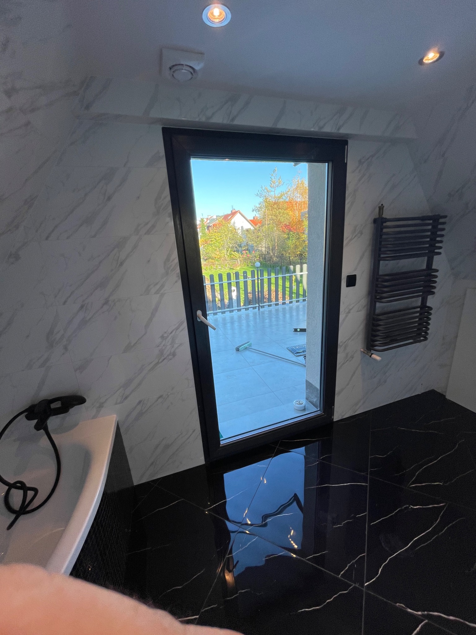 Nowoczesna łazienka z czarno-białymi marmurkowymi płytkami, widok na balkon przez czarne drzwi balkonowe i grzejnik drabinkowy na ścianie.