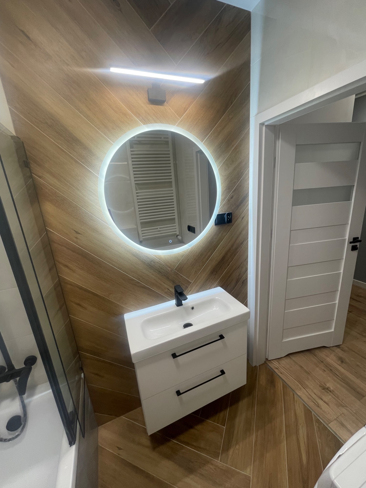 Nowoczesna łazienka z drewnopodobnymi płytkami ułożonymi w jodełkę na ścianie i podłodze, okrągłe lustro z podświetleniem LED nad białą szafką z umywalką i czarną armaturą, widoczna kabina...