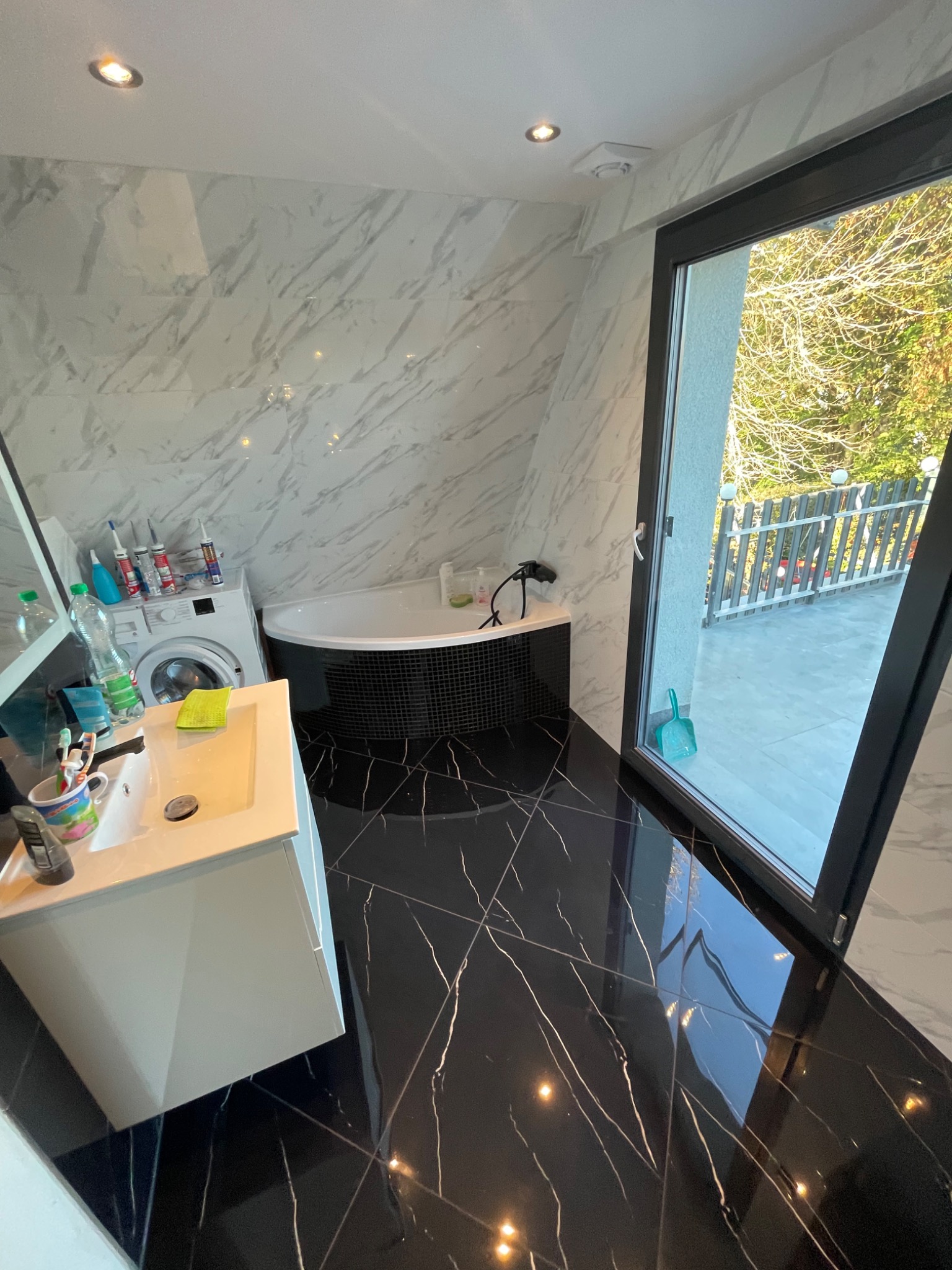 Nowoczesna łazienka z czarną marmurową podłogą, białą umywalką i wanną narożną, pralką oraz wyjściem na balkon z widokiem na drzewa.