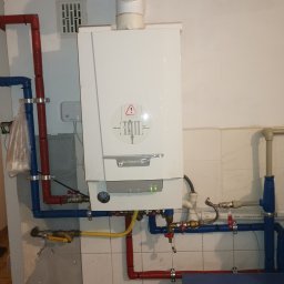 Instalacje sanitarne serwis urządzeń grzewczych - Biały piec gazowy marki De Dietrich zamontowany na ścianie z podłączonymi rurami w kolorach czerwonym i niebieskim, widoczne zawory i manometr, na dole fragment niebieskiego blatu z narzędziami...