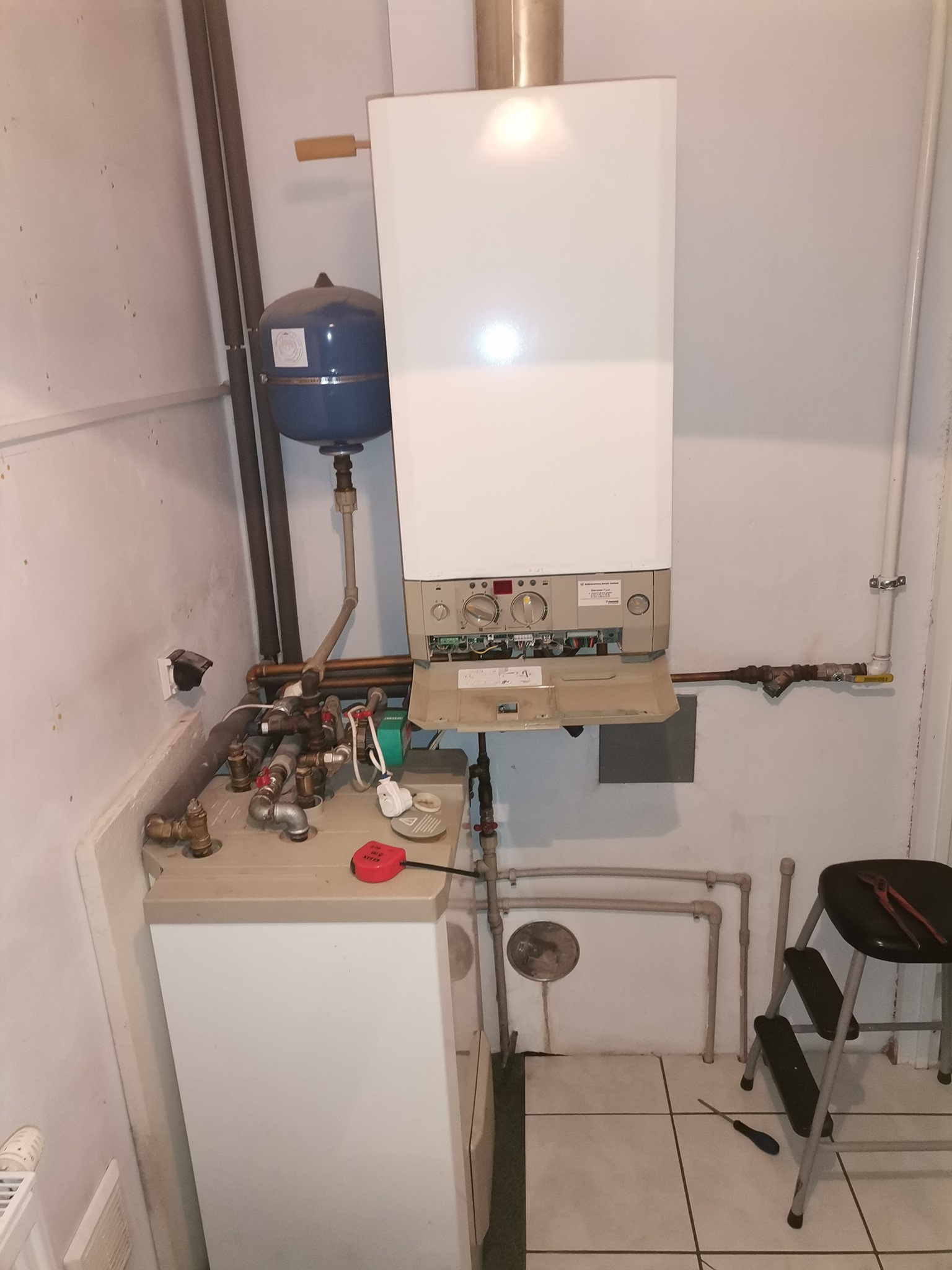 Biały piec gazowy zamontowany na ścianie w pomieszczeniu gospodarczym, widoczne rury, zbiornik wyrównawczy, drabinka z narzędziami i płytki na podłodze.