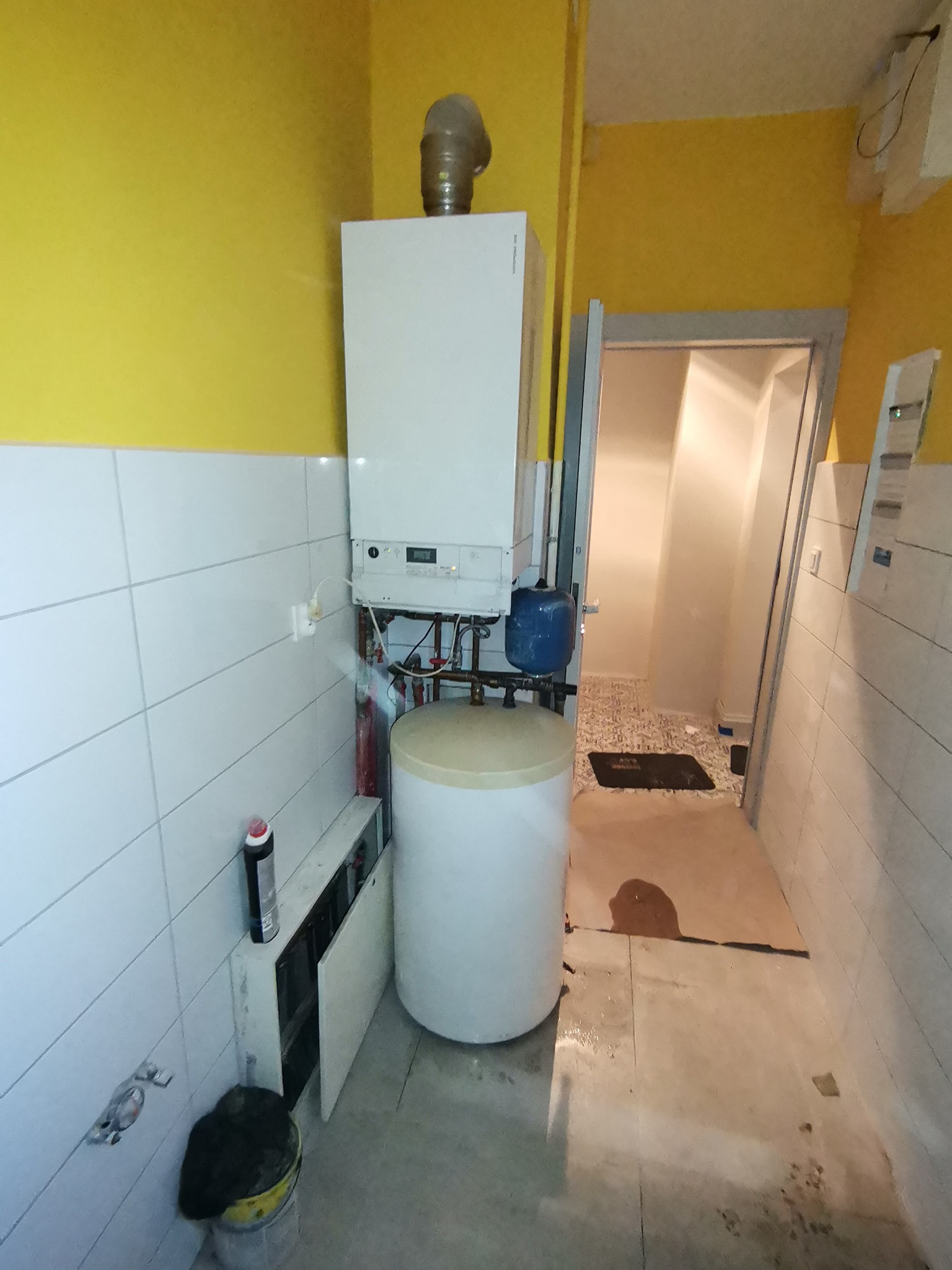 Biały piec centralnego ogrzewania z zasobnikiem ciepłej wody, rurami i niebieskim naczyniem wzbiorczym na tle żółtej ściany i białych płytek, widoczne także fragmenty instalacji grzewczej i otwarte...