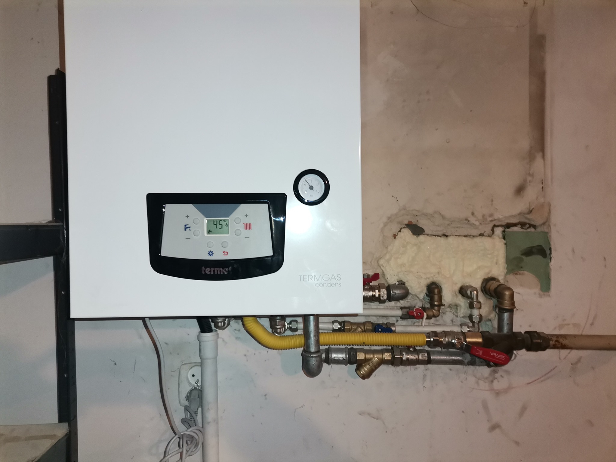 Biały piec gazowy dwufunkcyjny marki Termet z widocznymi przyłączami hydraulicznymi i gazowymi na tle ściany w surowym stanie.