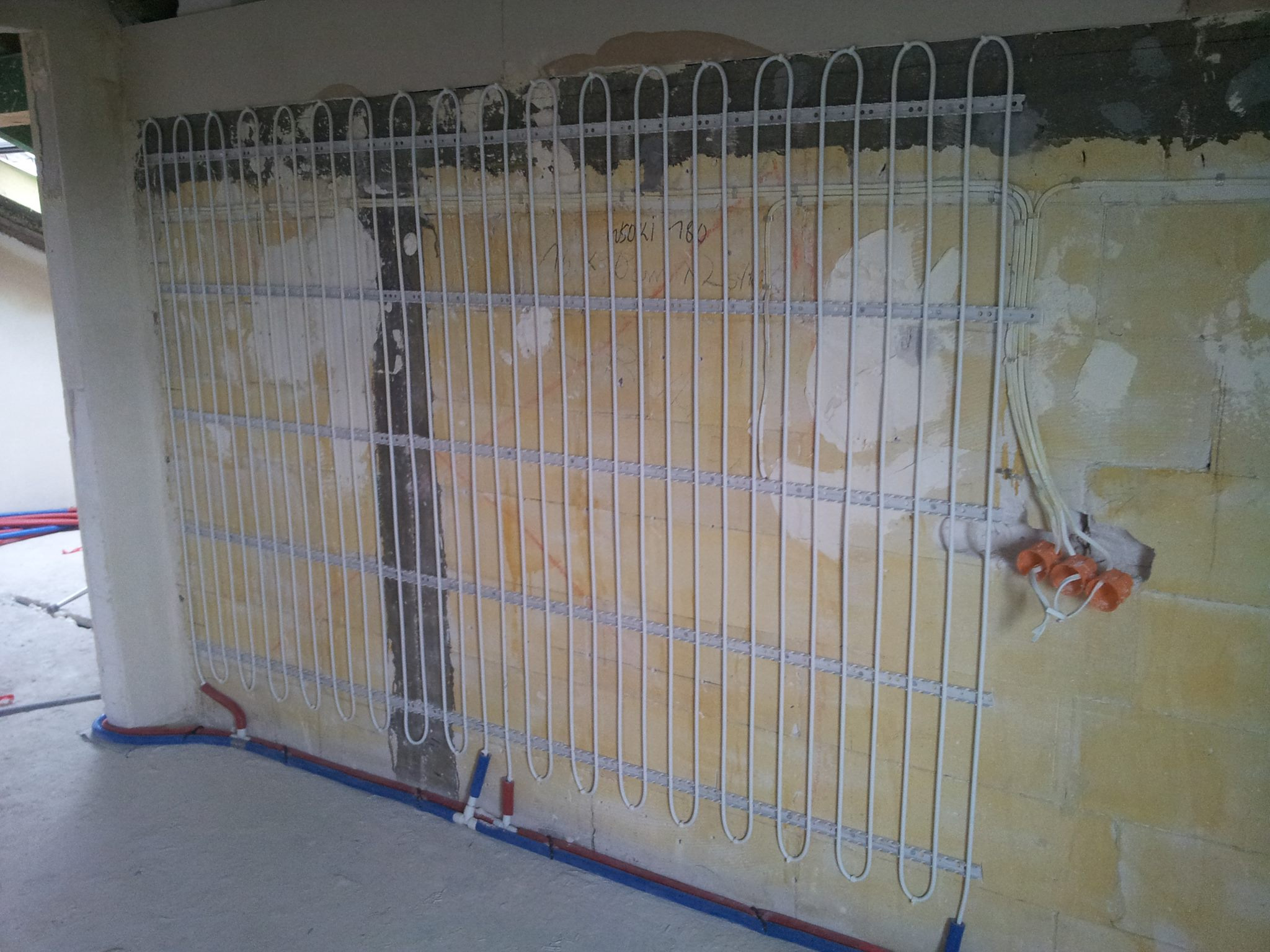 Montaż rur ogrzewania podłogowego na ścianie w trakcie realizacji w Warszawie, widoczne mocowania i fragment instalacji hydraulicznej.