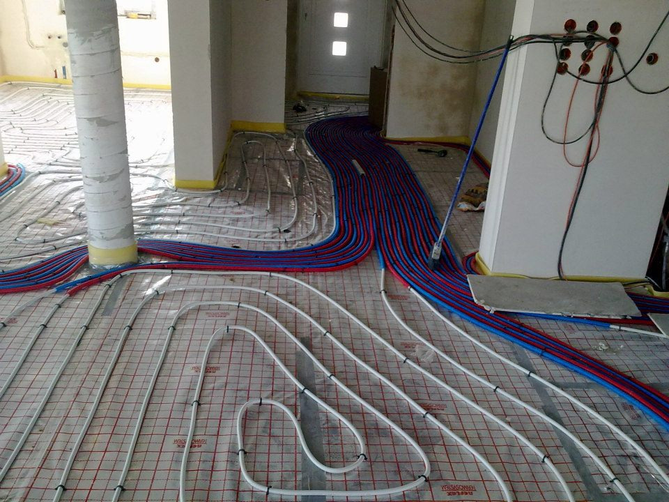 Instalacja ogrzewania podłogowego w trakcie montażu: rury grzewcze ułożone na folii z siatką, widoczne kable elektryczne w ścianie i przygotowane otwory w posadzce.