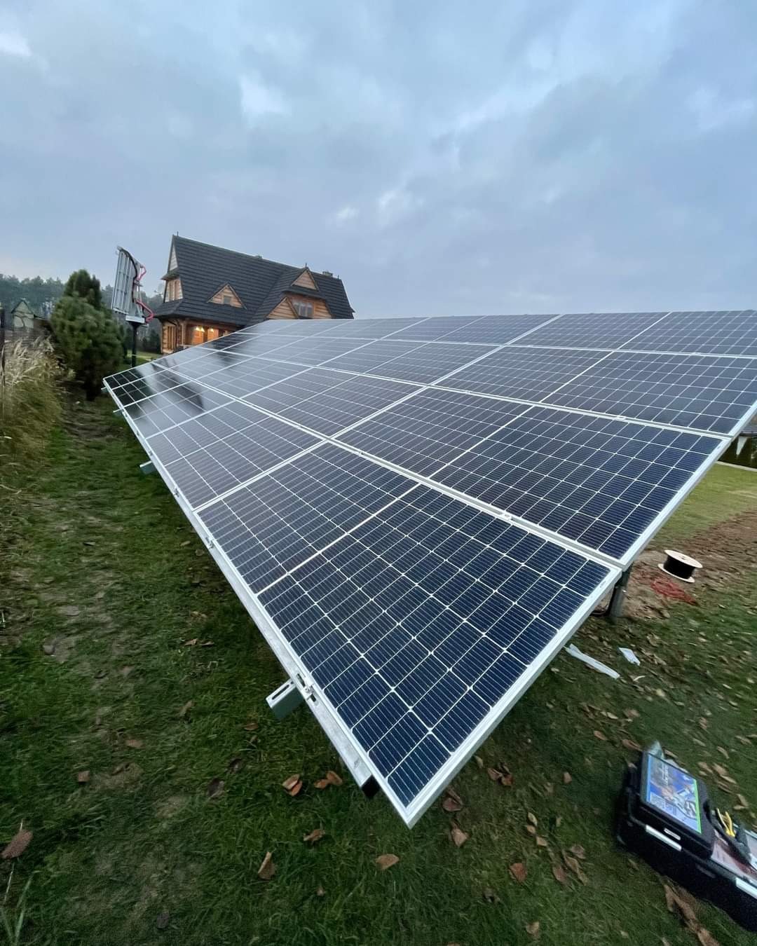 Instalacja paneli słonecznych na trawie przed domem z ciemnego drewna w pochmurny dzień.