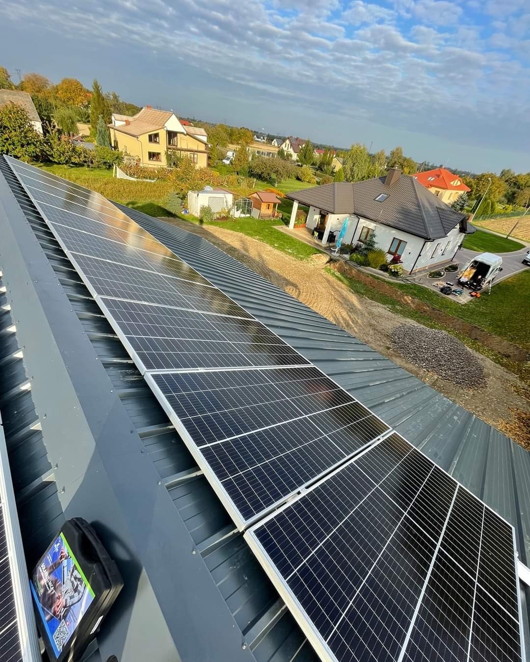 Panele słoneczne zamontowane na szarym, blaszanym dachu domu jednorodzinnego, widok z góry, w tle zabudowa mieszkalna i błękitne niebo z chmurami.