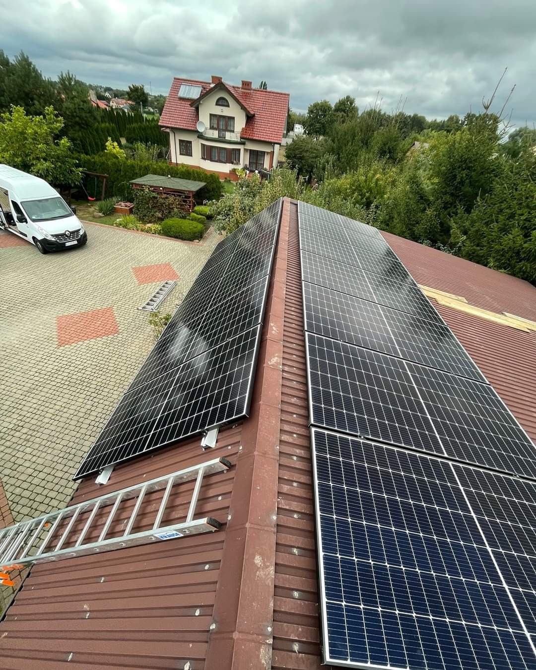 Montaż paneli słonecznych na dachu domu jednorodzinnego z blachy trapezowej; widoczna drabina oparta o dach i biały samochód dostawczy w tle.