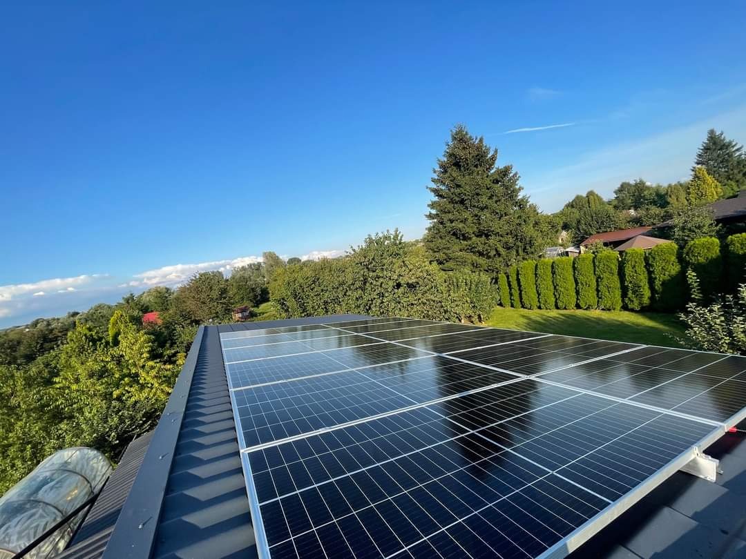 Panele słoneczne na ciemnym dachu z widokiem na zielone drzewa i błękitne niebo z delikatnymi chmurami.