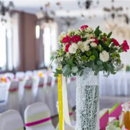 Dekoracja stołu z wysokim wazonem owiniętym białą siatką, wypełnionym bukietem z białych, różowych i zielonych kwiatów, z żółtymi wstążkami, na tle sali bankietowej z nakrytymi stołami i krzesłami.