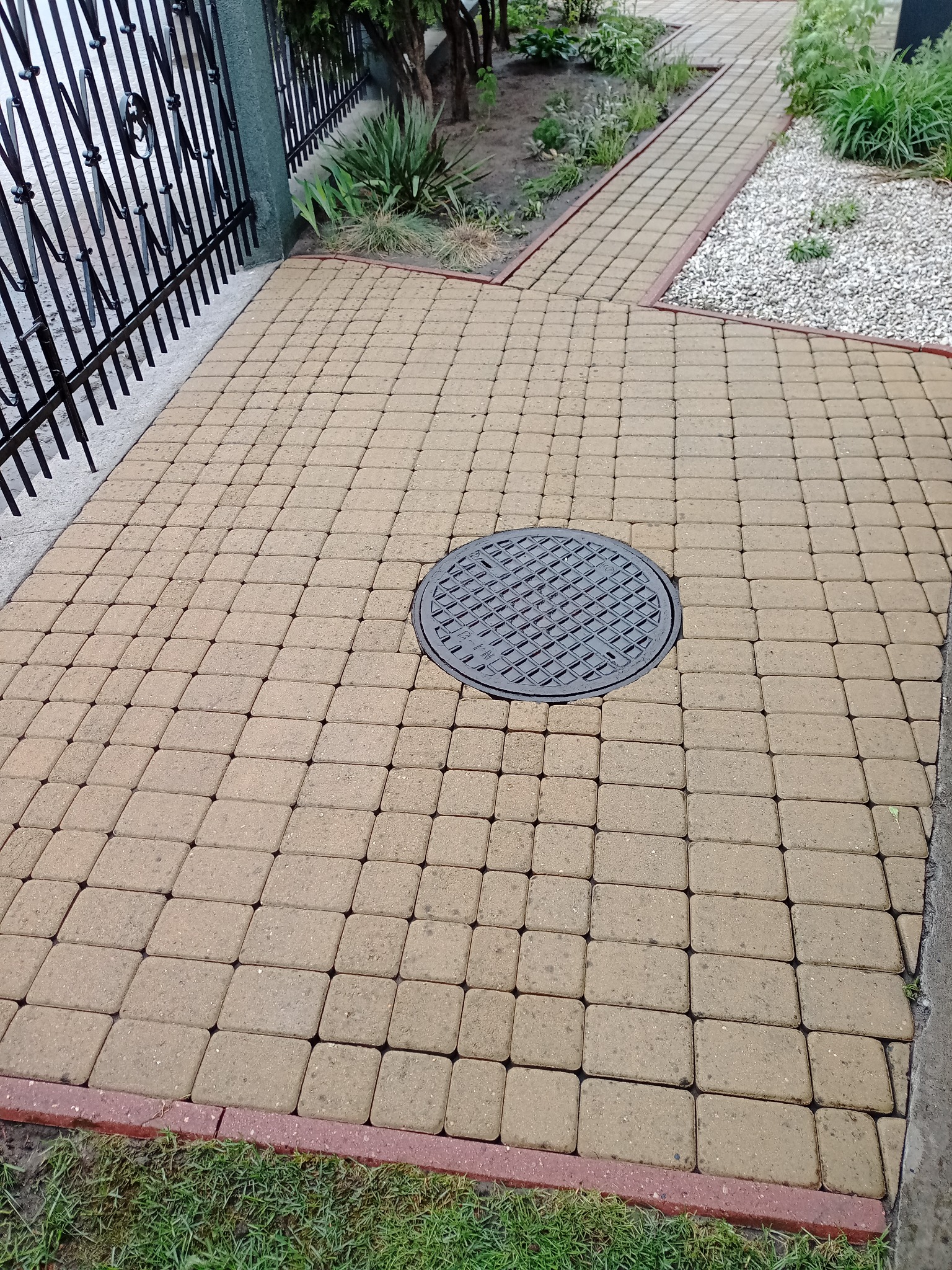 Ułożona kostka brukowa w odcieniach beżu, z widocznym włazem kanalizacyjnym pośrodku, obrzeżona czerwoną kostką, z fragmentem metalowego ogrodzenia w tle.