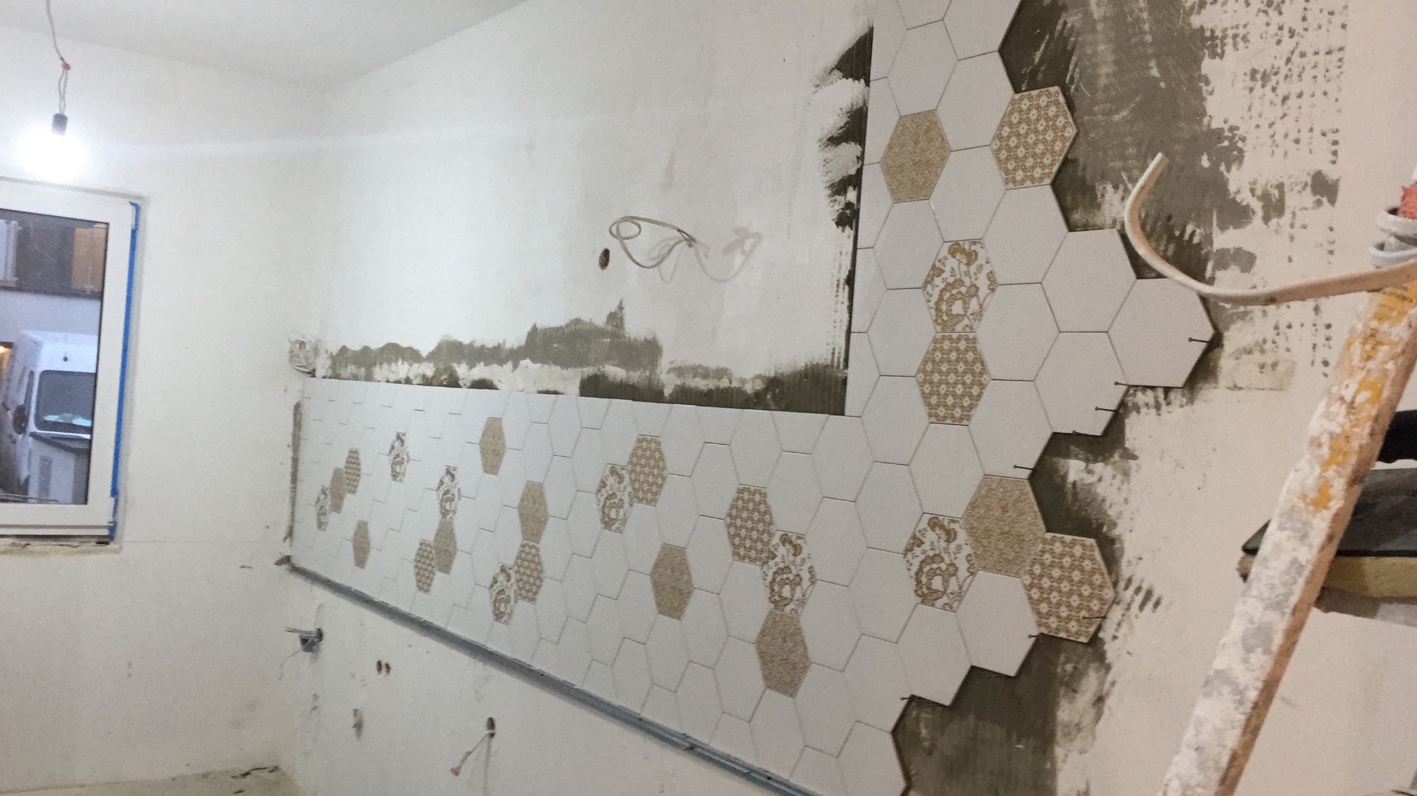Montaż heksagonalnych płytek z wzorem na ścianie w trakcie prac, widoczne ślady zaprawy i drabina oparta o ścianę, okno z widokiem na samochód dostawczy.