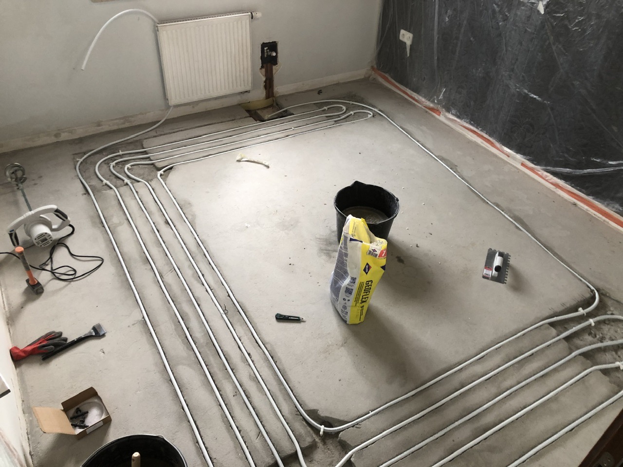 Rozłożone rury ogrzewania podłogowego w trakcie montażu, widoczny worek z zaprawą, narzędzia i zabezpieczona folią ściana.