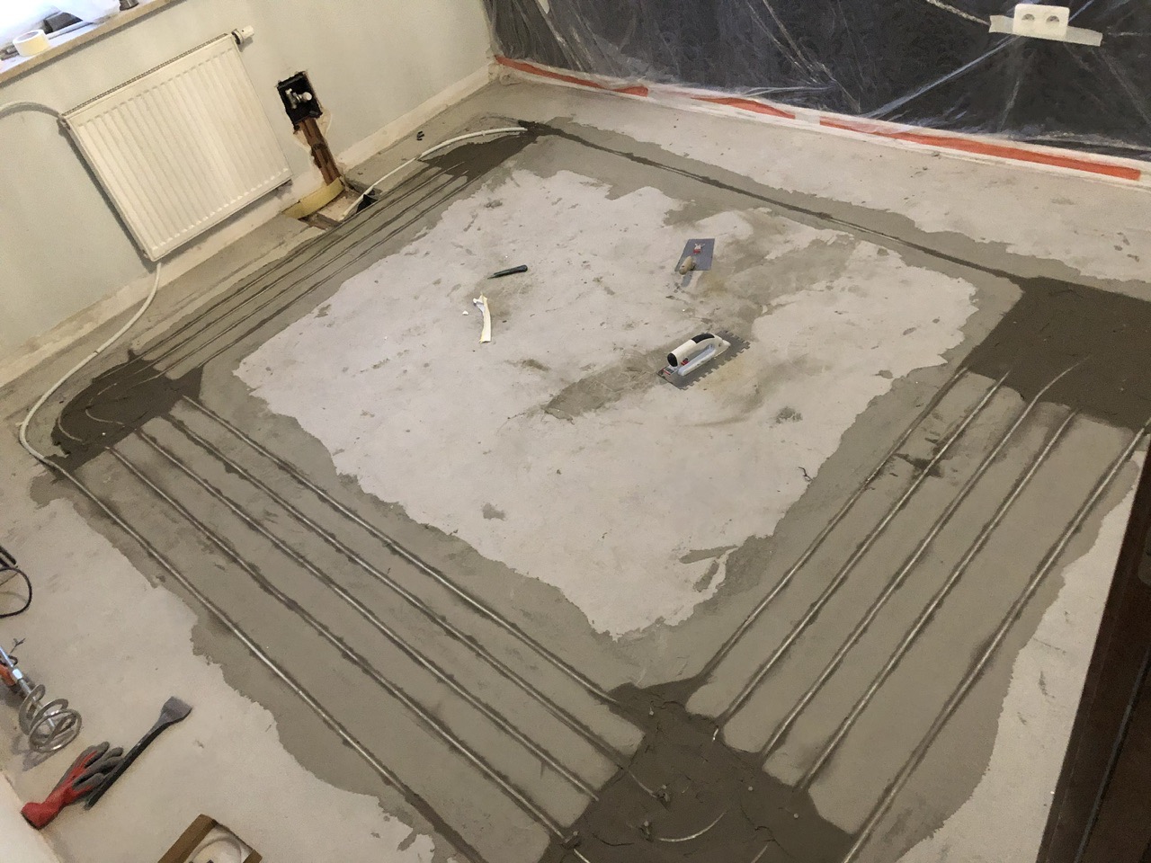 Montaż ogrzewania podłogowego: rury grzewcze ułożone w pętle i zalewane wylewką, widoczne narzędzia glazurnicze i instalacyjne.