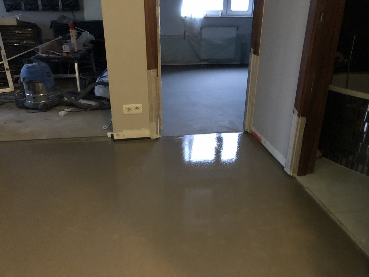 Świeżo wylana, samopoziomująca wylewka podłogowa w pomieszczeniu, widoczne narzędzia i odkurzacz przemysłowy w tle.