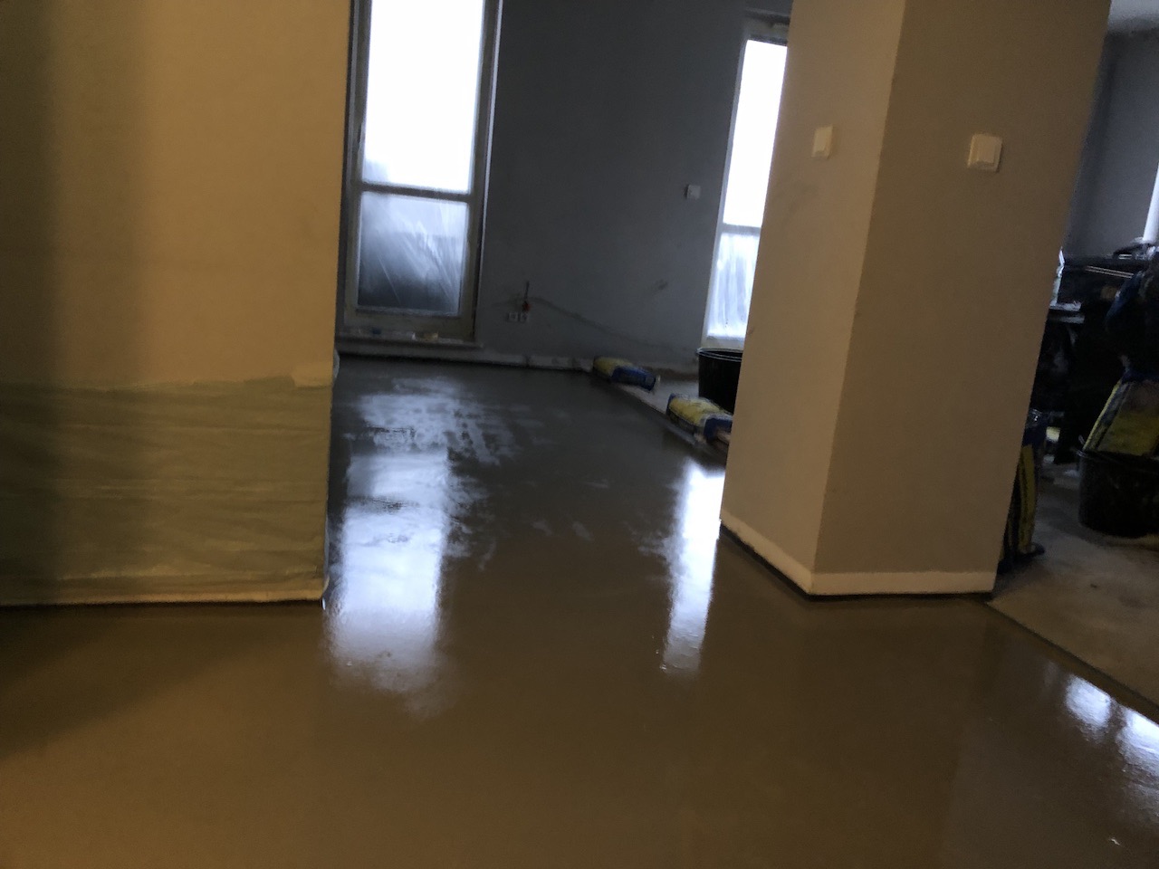 Wylewka samopoziomująca na podłodze w trakcie prac wykończeniowych, widoczne okna i filary, worki z materiałem budowlanym w tle.