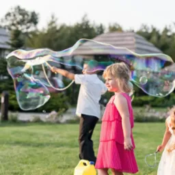 Animator dmucha ogromne bańki mydlane dla dzieci podczas imprezy plenerowej, widoczna dziewczynka w różowej sukience i chłopiec w tle.