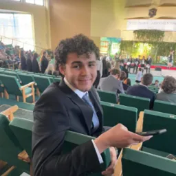 Młody mężczyzna w garniturze siedzi na widowni podczas uroczystości, trzyma telefon w dłoni, w tle scena z napisem 'Pożegnanie Absolwent&oacute;w 2023'