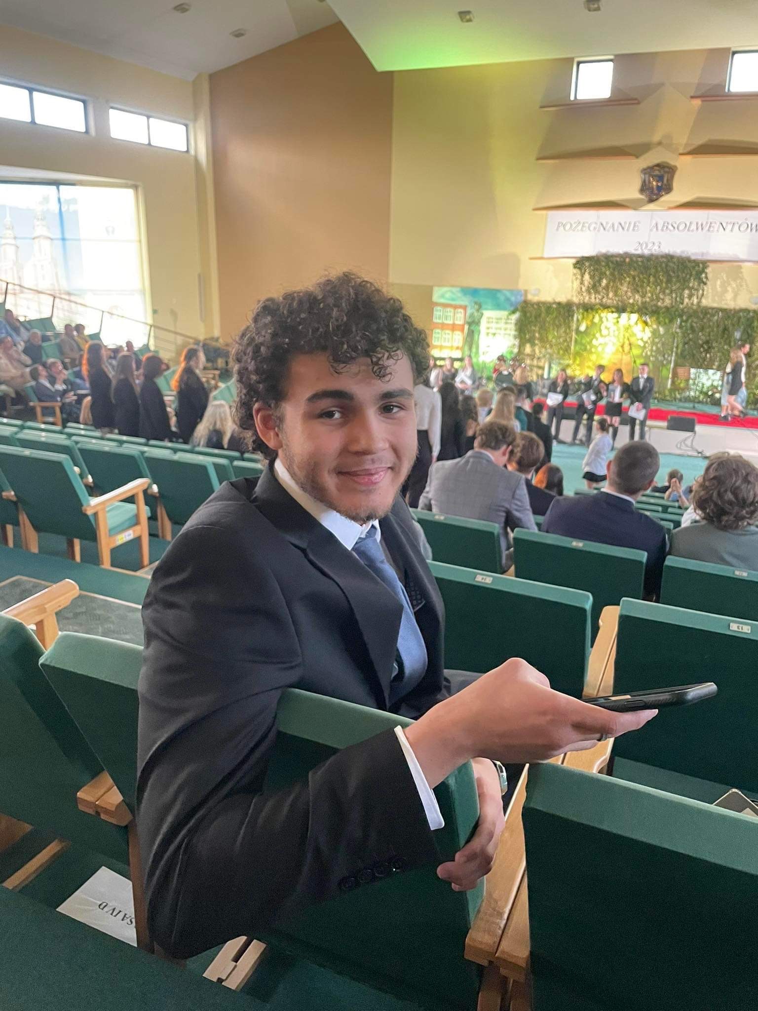 Młody mężczyzna w garniturze siedzi na widowni podczas uroczystości, trzyma telefon w dłoni, w tle scena z napisem 'Pożegnanie Absolwentów 2023'