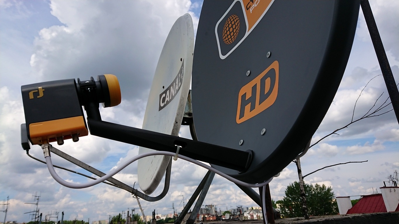 Dwie anteny satelitarne zamontowane na dachu budynku, jedna z logo 'CANAL+', druga z oznaczeniem 'HD', widok na pochmurne niebo i zabudowę miejską w tle.