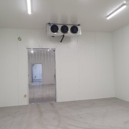 Sigma Chłodnictwo-serwis - Wnętrze chłodni z trzema wentylatorami zamontowanymi na suficie, widoczna kurtyna paskowa przy wejściu i wzorzysta podłoga. Białe ściany i jasne oświetlenie sufitowe.