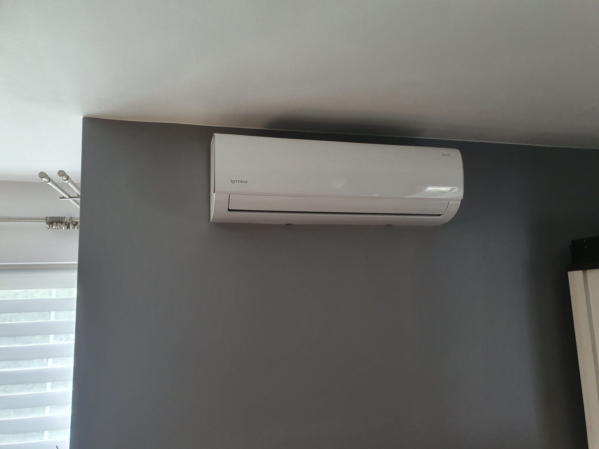 Biały klimatyzator marki Rotenso zamontowany na szarej ścianie w pomieszczeniu z białym sufitem i żaluzjami.