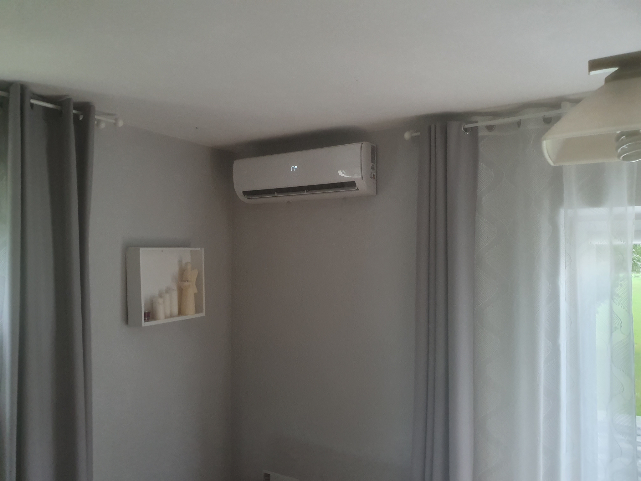 Biały klimatyzator ścienny z wyświetlaną temperaturą 17 stopni Celsjusza, zamontowany w szarym pokoju z zasłonami i dekoracją ścienną.