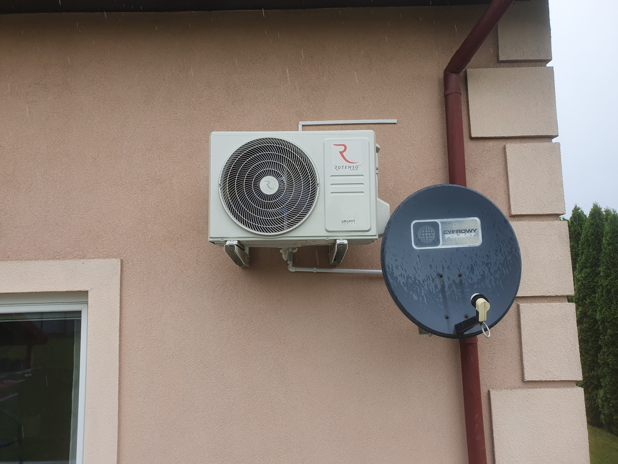 Biała jednostka zewnętrzna klimatyzatora Rotenso zamontowana na elewacji budynku obok anteny satelitarnej, widoczne krople deszczu.