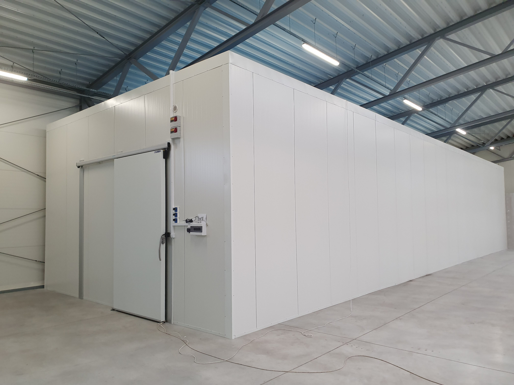 Duża, biała chłodnia przemysłowa z przesuwnymi drzwiami w hali magazynowej, widoczne panele ścienne i sufit z oświetleniem.