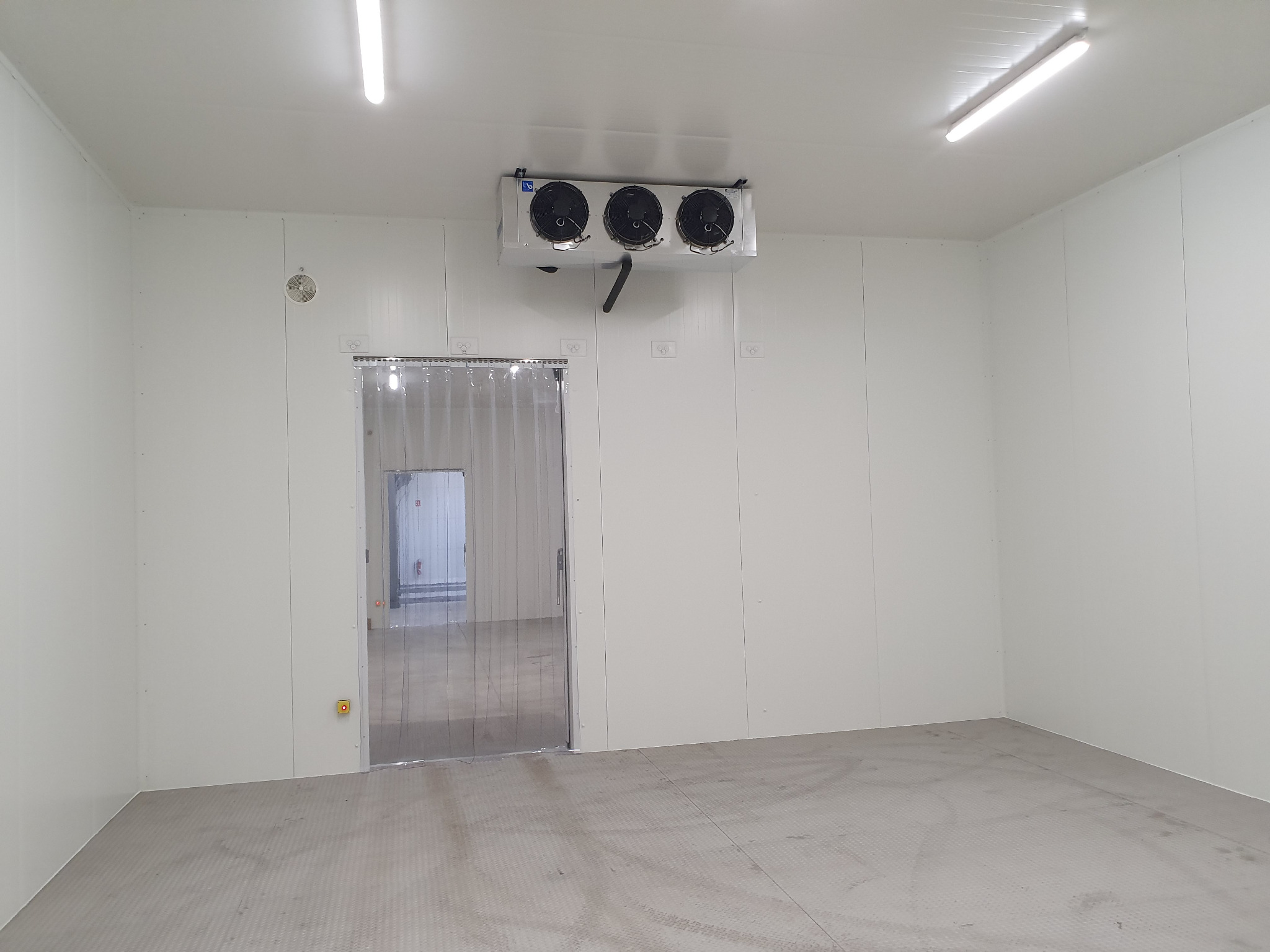 Wnętrze chłodni z trzema wentylatorami zamontowanymi na suficie, widoczna kurtyna paskowa przy wejściu i wzorzysta podłoga. Białe ściany i jasne oświetlenie sufitowe.