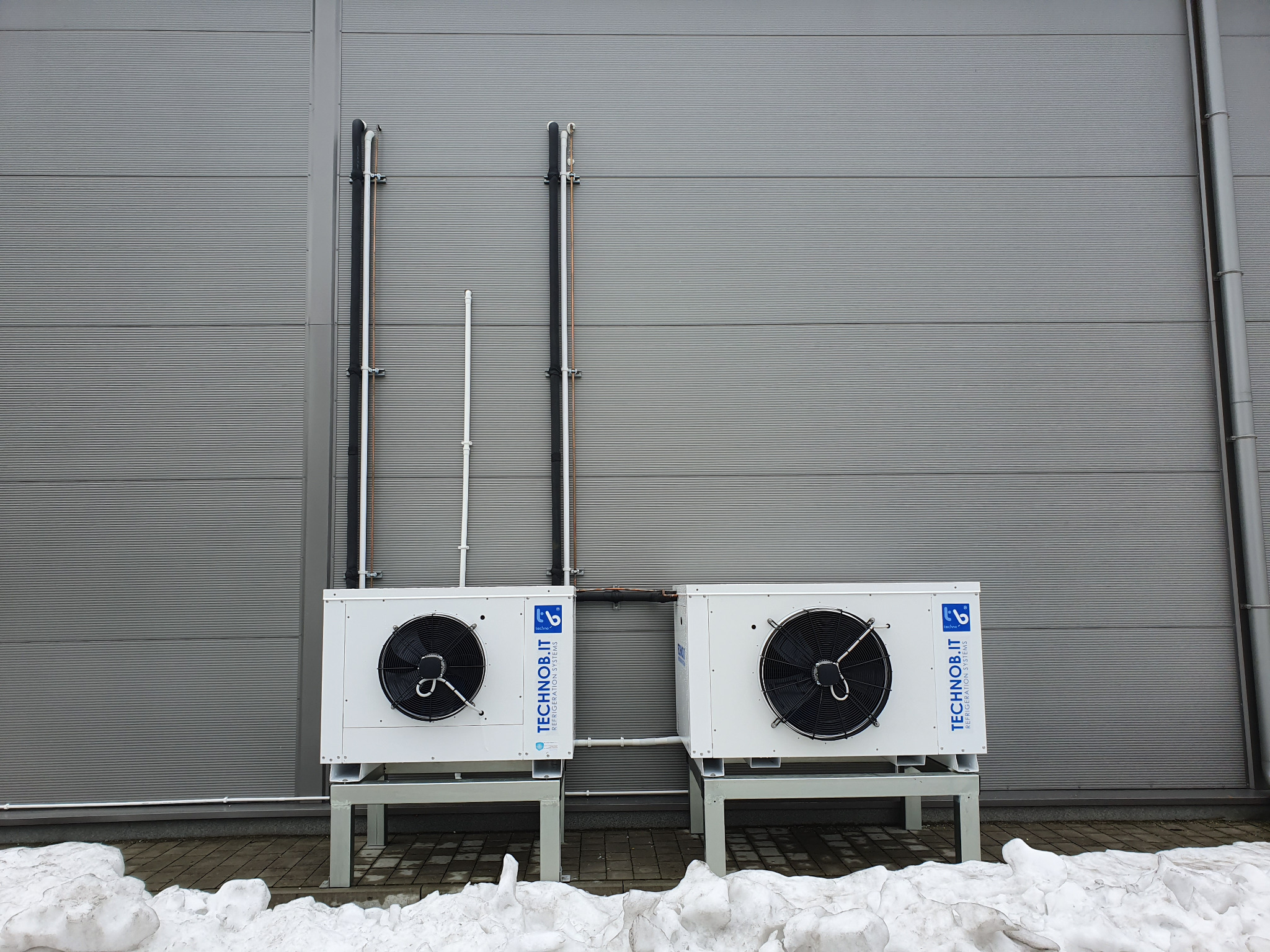 Dwa białe agregaty chłodnicze TECHNOb.IT na metalowych podstawach, zamontowane na zewnątrz budynku z szarej blachy trapezowej, widoczne rury instalacyjne i resztki topniejącego śniegu.