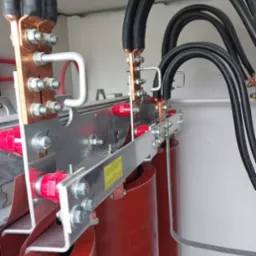 Szczegółowe ujęcie fragmentu transformatora z widocznymi miedzianymi szynami, czarnymi kablami, aluminiowymi elementami konstrukcyjnymi i czerwonymi izolatorami.