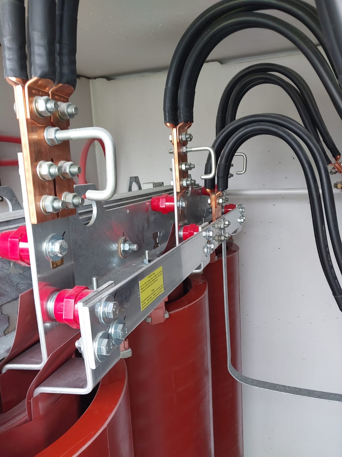 Szczegółowe ujęcie fragmentu transformatora z widocznymi miedzianymi szynami, czarnymi kablami, aluminiowymi elementami konstrukcyjnymi i czerwonymi izolatorami.