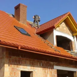 Montaż blachodachówki w kolorze ceglastym na nowym domu, widoczne okno dachowe, komin z cegły, rynny oraz dekarz w kombinezonie na dachu, w tle błękitne niebo.