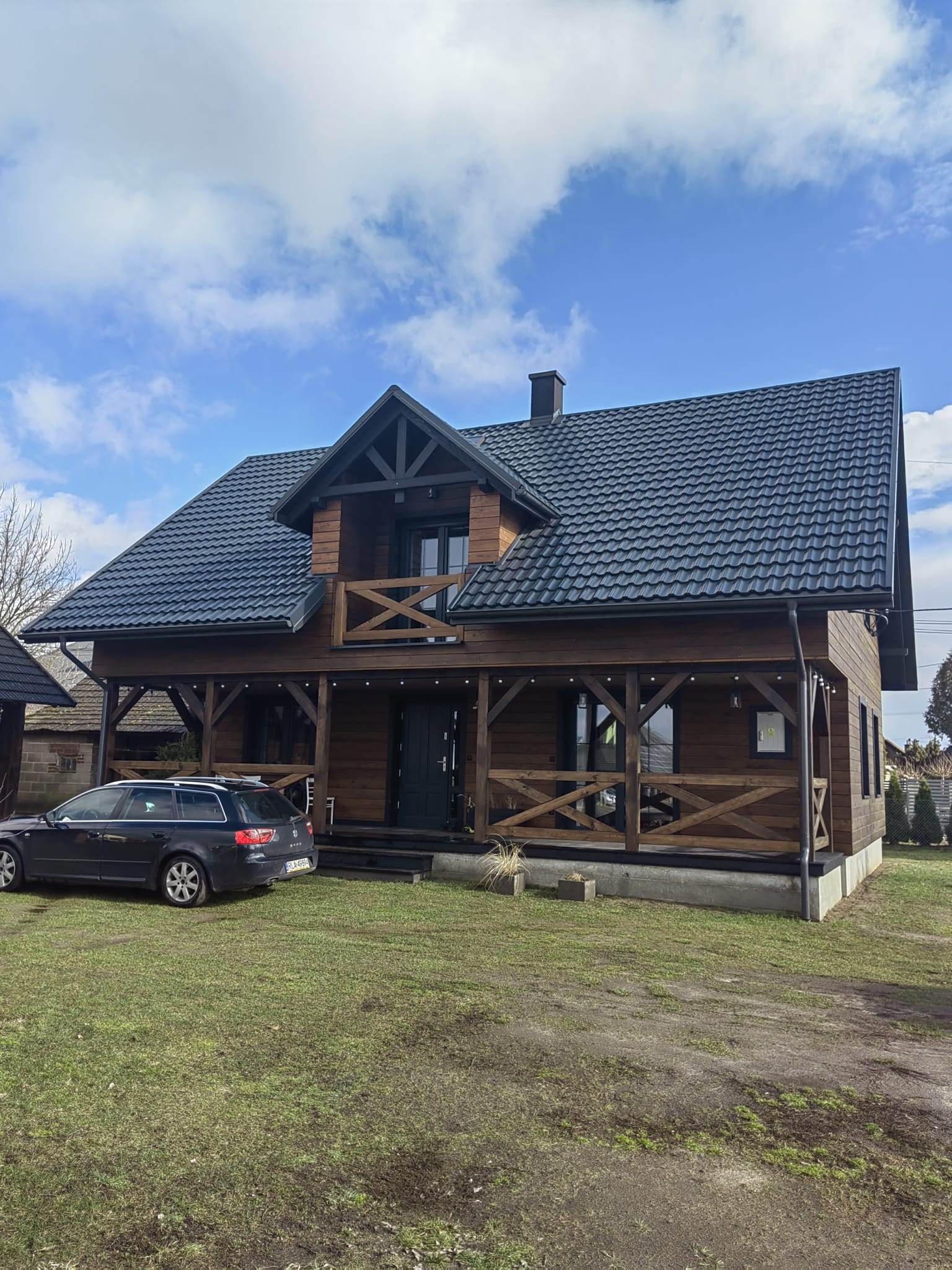 Nowy dom z ciemnym dachem z blachy trapezowej, drewnianą elewacją i werandą z ozdobnymi krzyżulcami, na zadbanym trawniku, z zaparkowanym samochodem osobowym.