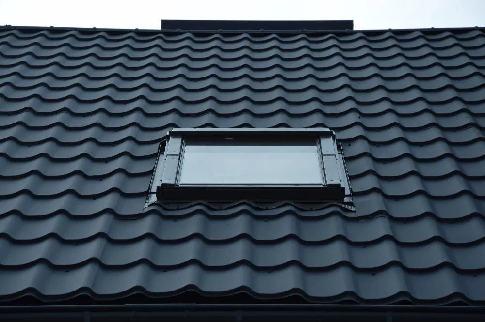 Ciemna dachówka ceramiczna z oknem dachowym, widoczne faliste ułożenie dachówek i czarne obramowanie okna.