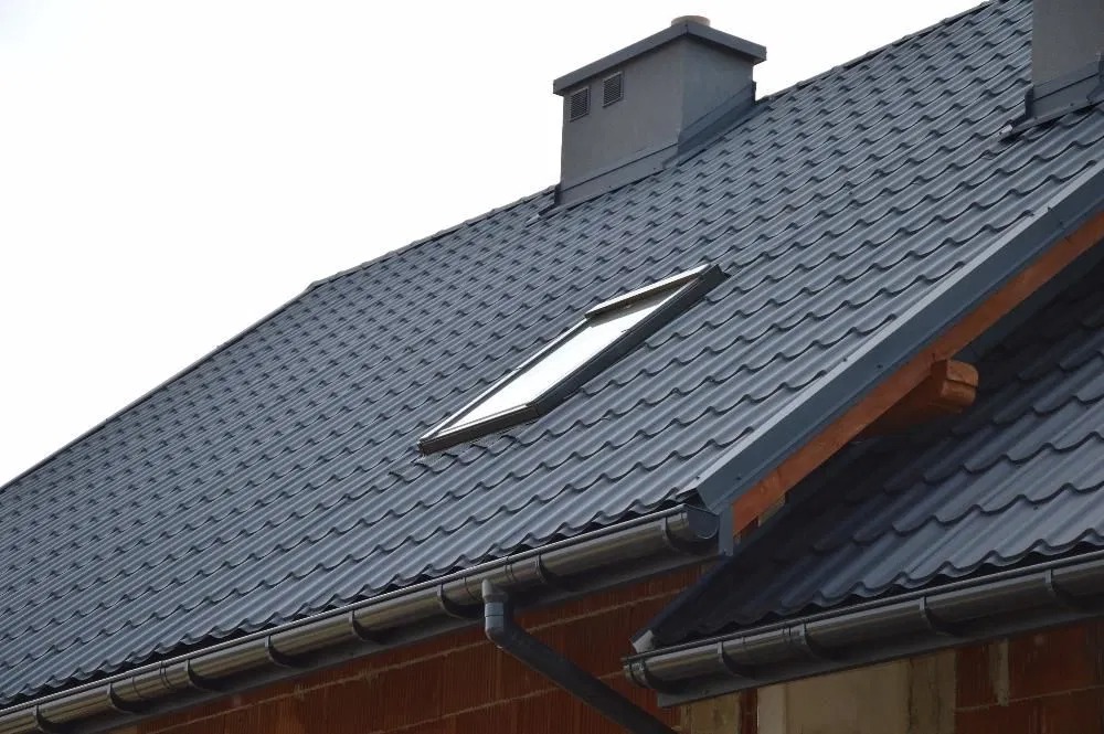 Szary dach z blachodachówki z oknem dachowym i szarym kominem, widoczne rynny i fragment ściany z cegły.