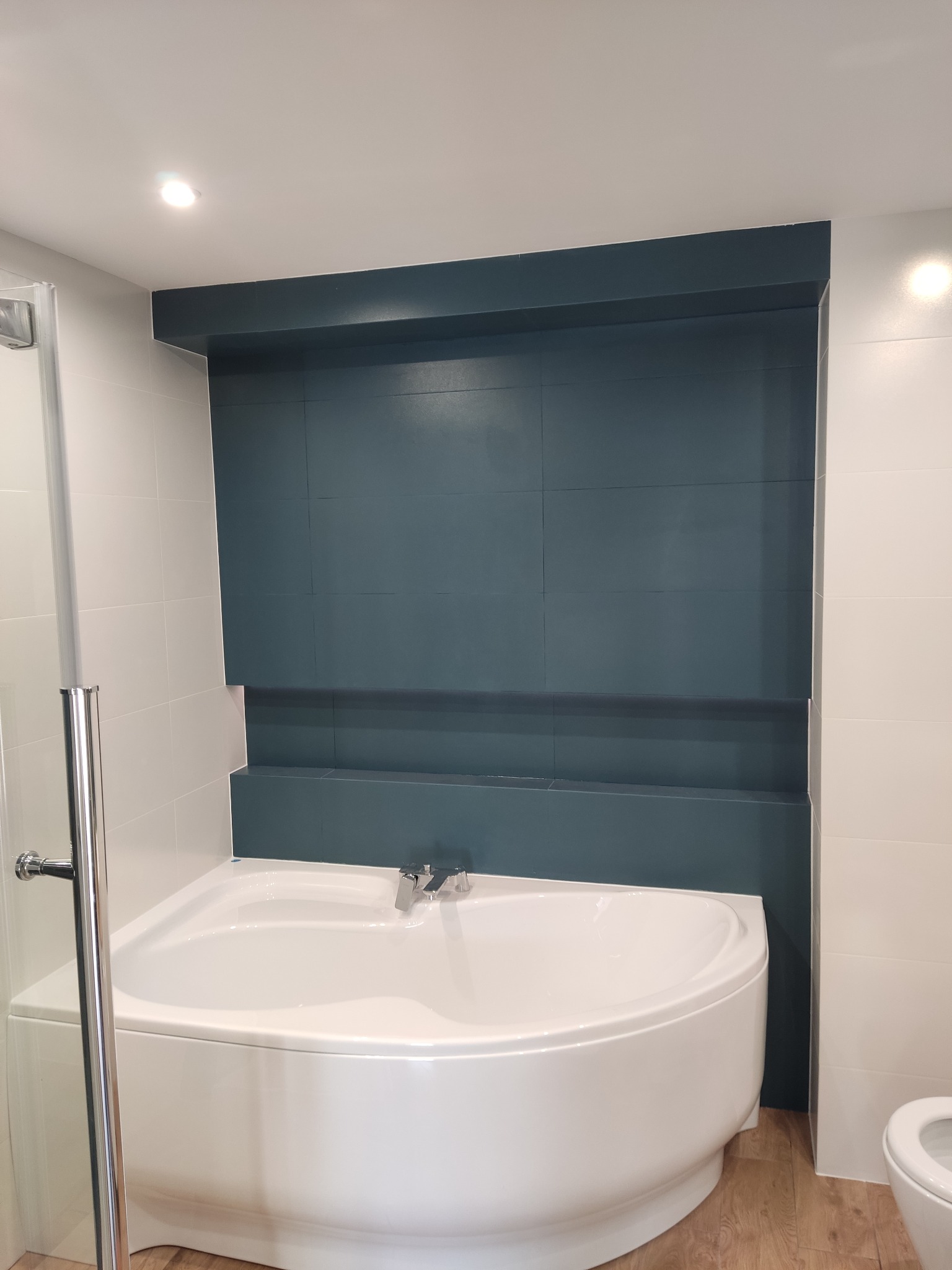 Narożna wanna w wykończonej łazience z ciemnoturkusową ścianą w zabudowie i jasnymi płytkami oraz drewnopodobną podłogą, widoczna kabina prysznicowa z chromowanymi elementami.