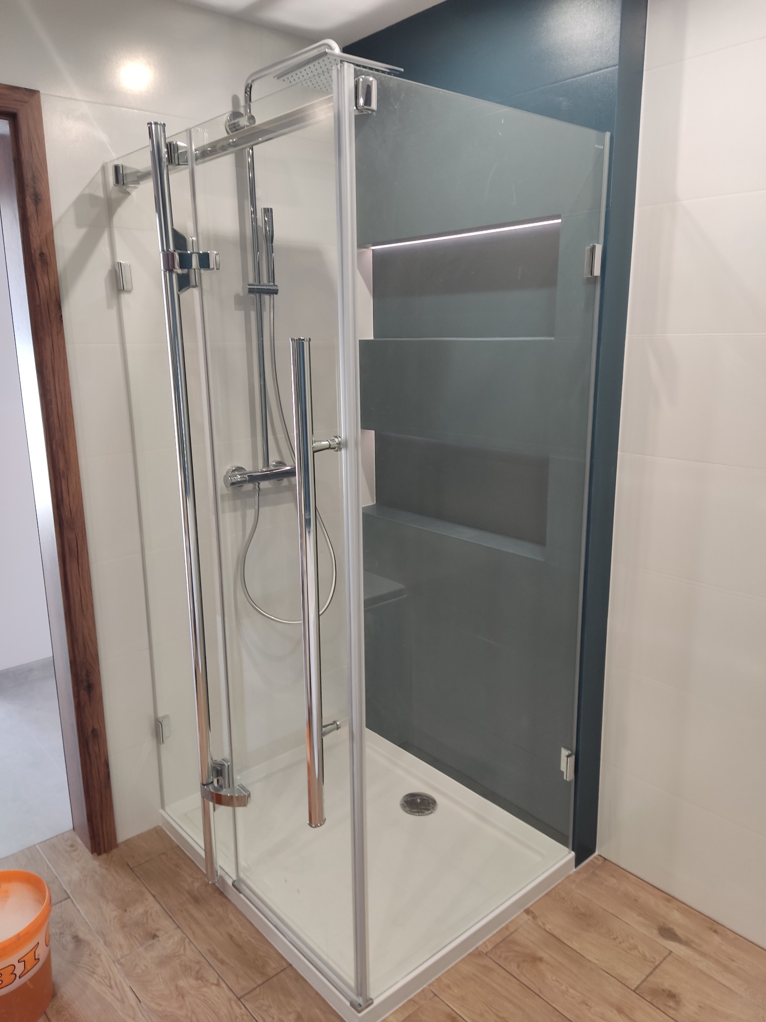 Nowoczesna kabina prysznicowa z chromowanymi elementami, szklanymi drzwiami i ciemną ścianą w tle, widok na drewnopodobną podłogę i fragment drzwi wejściowych.