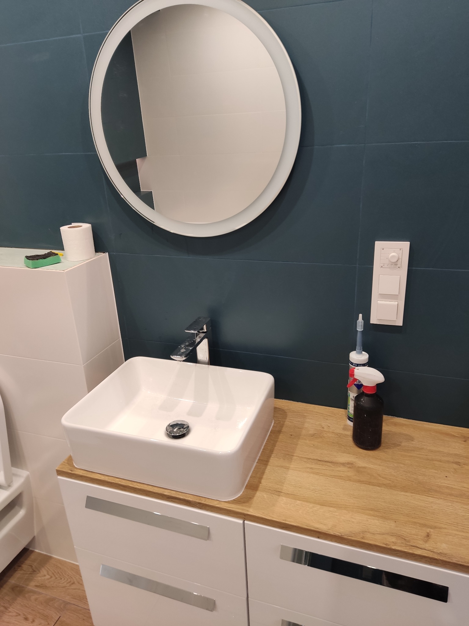 Nowoczesna łazienka z ciemnymi płytkami, okrągłym lustrem z podświetleniem LED, kwadratową umywalką nablatową, drewnianym blatem i białymi szafkami z podłużnymi uchwytami, widoczne akcesoria...