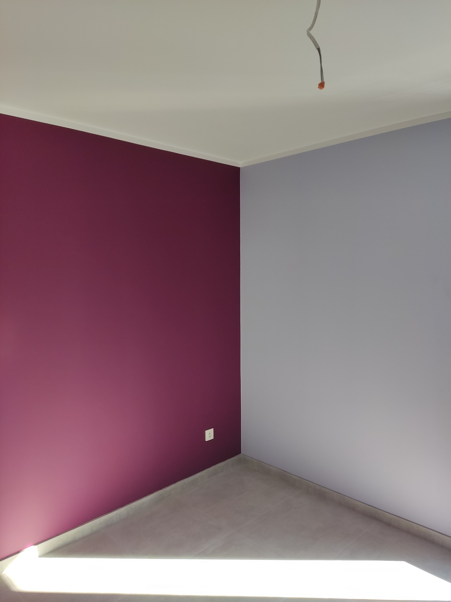 Pomieszczenie po malowaniu ścian na dwa kolory: purpurowy i szary, z widocznym gniazdkiem elektrycznym i wystającym przewodem z sufitu.