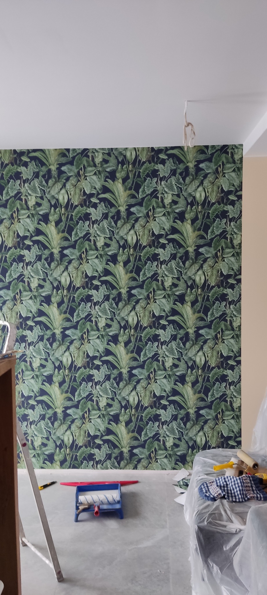 Ściana pokryta ciemną tapetą w tropikalne liście, widoczny wałek do tapetowania, drabina i zabezpieczone folią meble