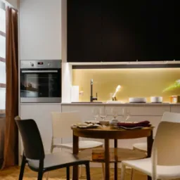 Nowoczesna, jasna kuchnia z czarnymi szafkami górnymi, okrągłym drewnianym stołem z nakryciem i czterema krzesłami, oraz dekoracjami na półkach i blacie.