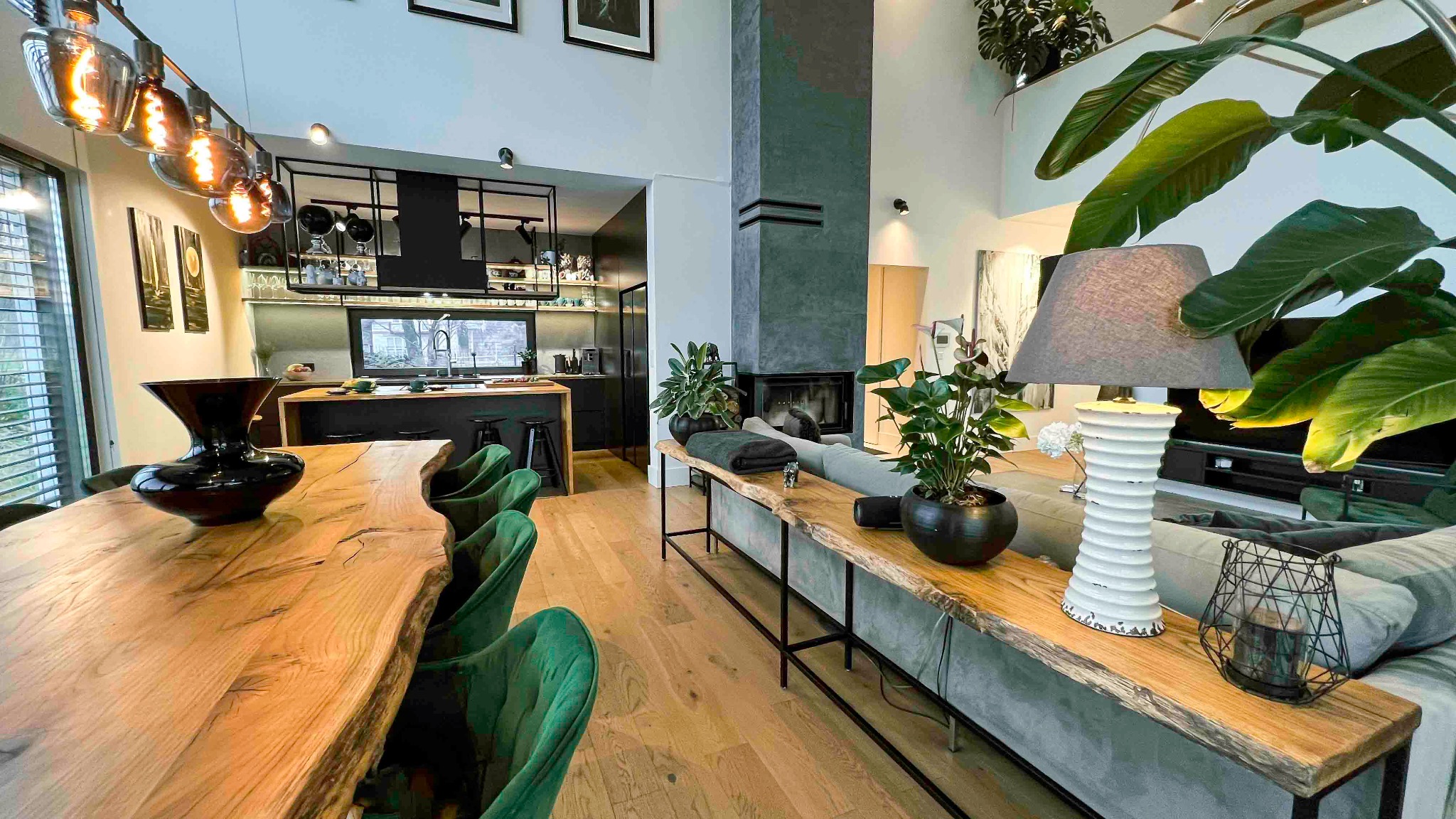 Przestronne wnętrze domu z otwartą kuchnią, jadalnią z drewnianym stołem z surowymi krawędziami, zielonymi krzesłami i salonem z kominkiem oraz roślinami doniczkowymi.