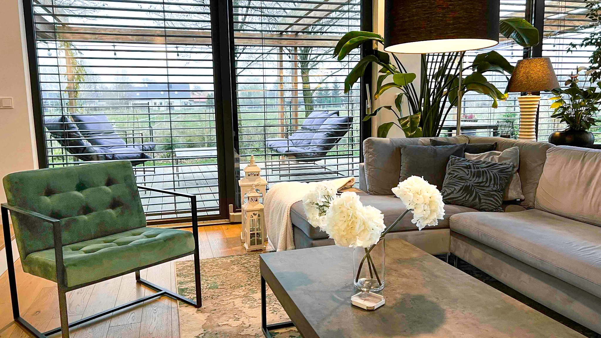 Nowoczesny salon z zielonym fotelem, szarą sofą, kwiatami w wazonie na betonowym stoliku kawowym i widokiem na ogród przez okno z żaluzjami.