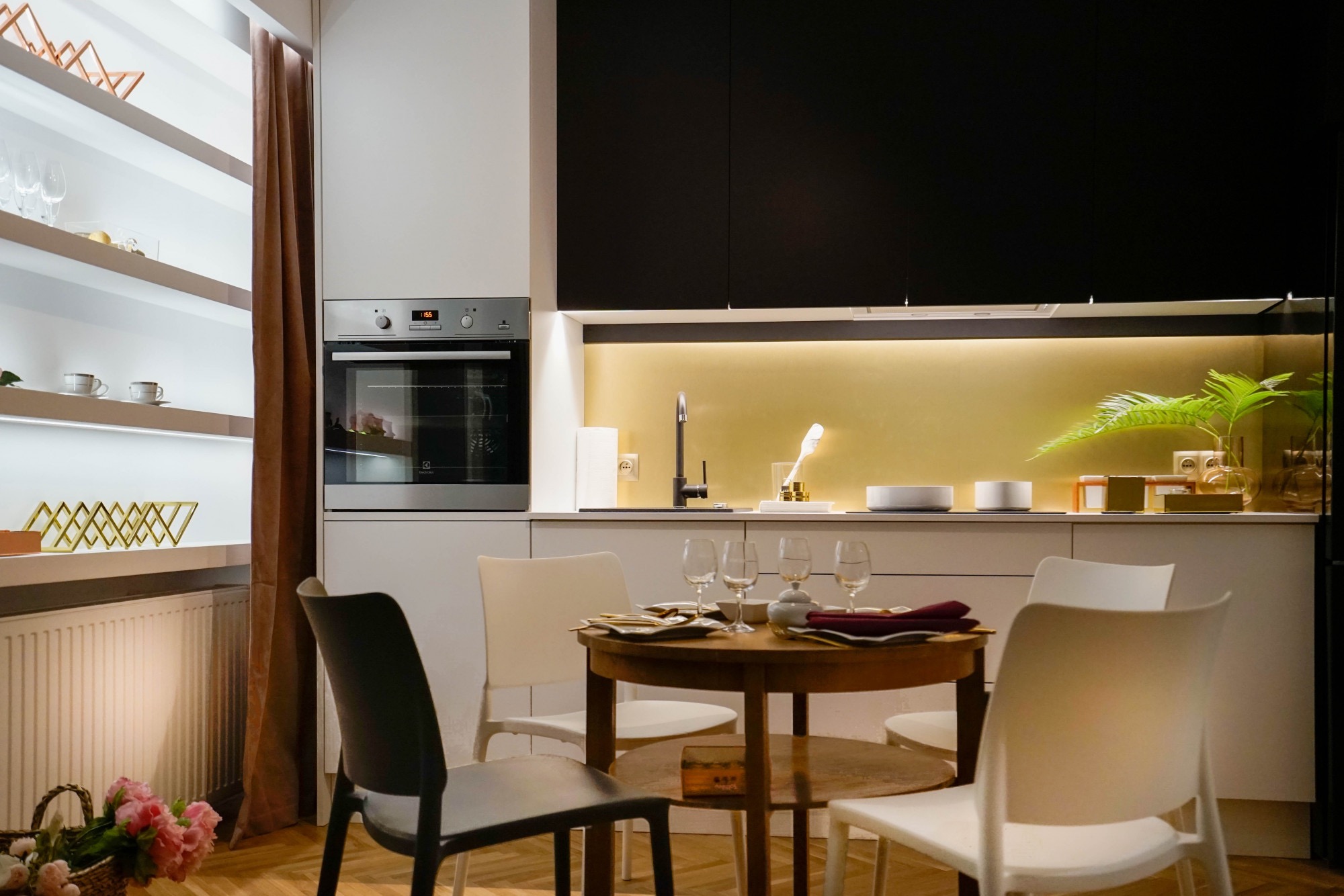 Nowoczesna, jasna kuchnia z czarnymi szafkami górnymi, okrągłym drewnianym stołem z nakryciem i czterema krzesłami, oraz dekoracjami na półkach i blacie.