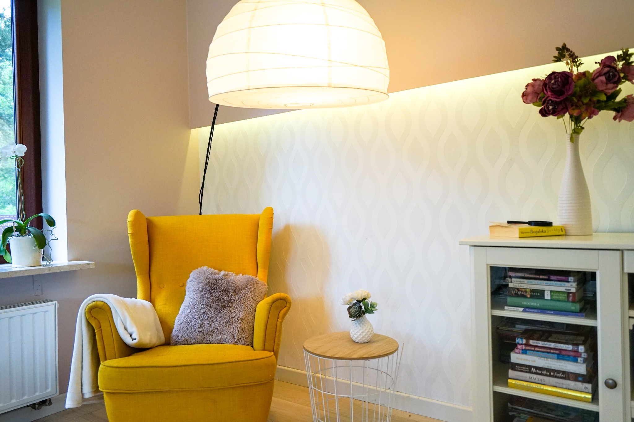 Żółty fotel uszak z poduszką i kocem w jasnym pokoju z kwiatami w wazonie i lampą wiszącą