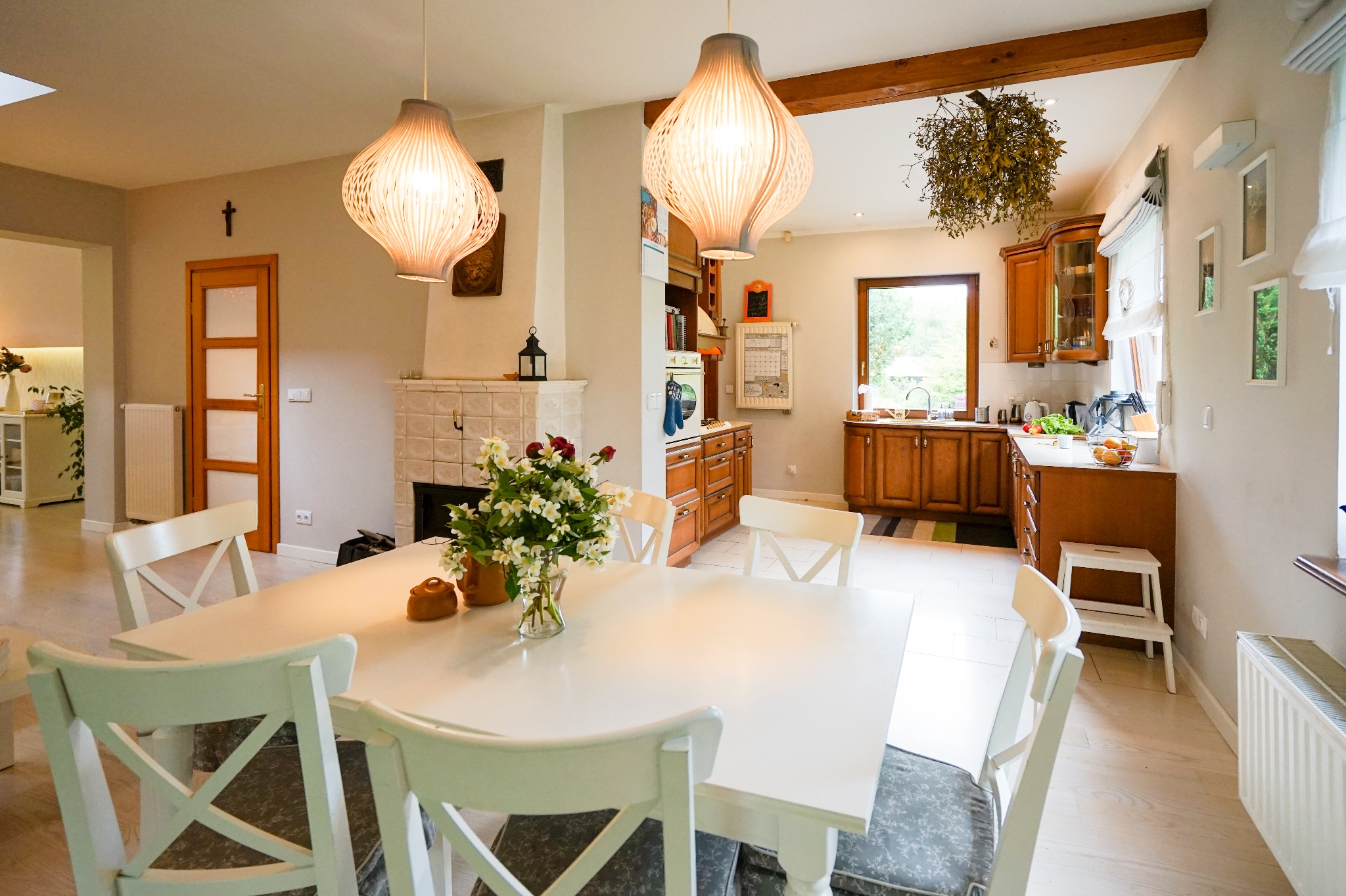 Jasne wnętrze jadalni z białym stołem i krzesłami, widok na kuchnię z drewnianymi szafkami, ozdobne lampy wiszące i kominek z kaflami.