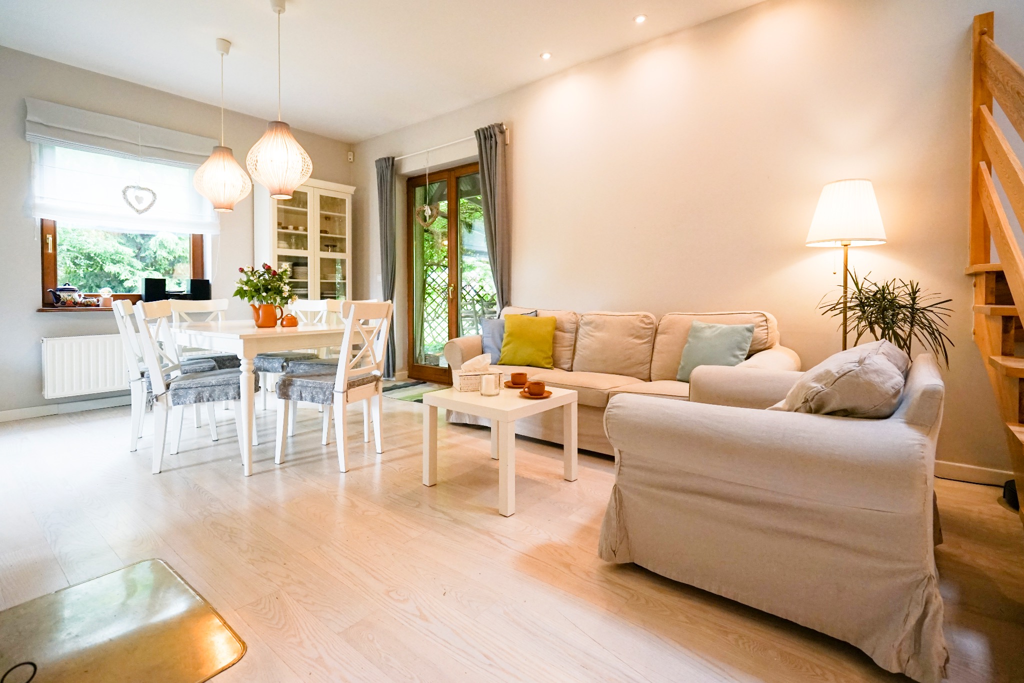 Jasne, przestronne wnętrze salonu z drewnianymi schodami, białym stołem i krzesłami, beżową sofą z kolorowymi poduszkami oraz lampą podłogową, oświetlone naturalnym światłem wpadającym przez okno.